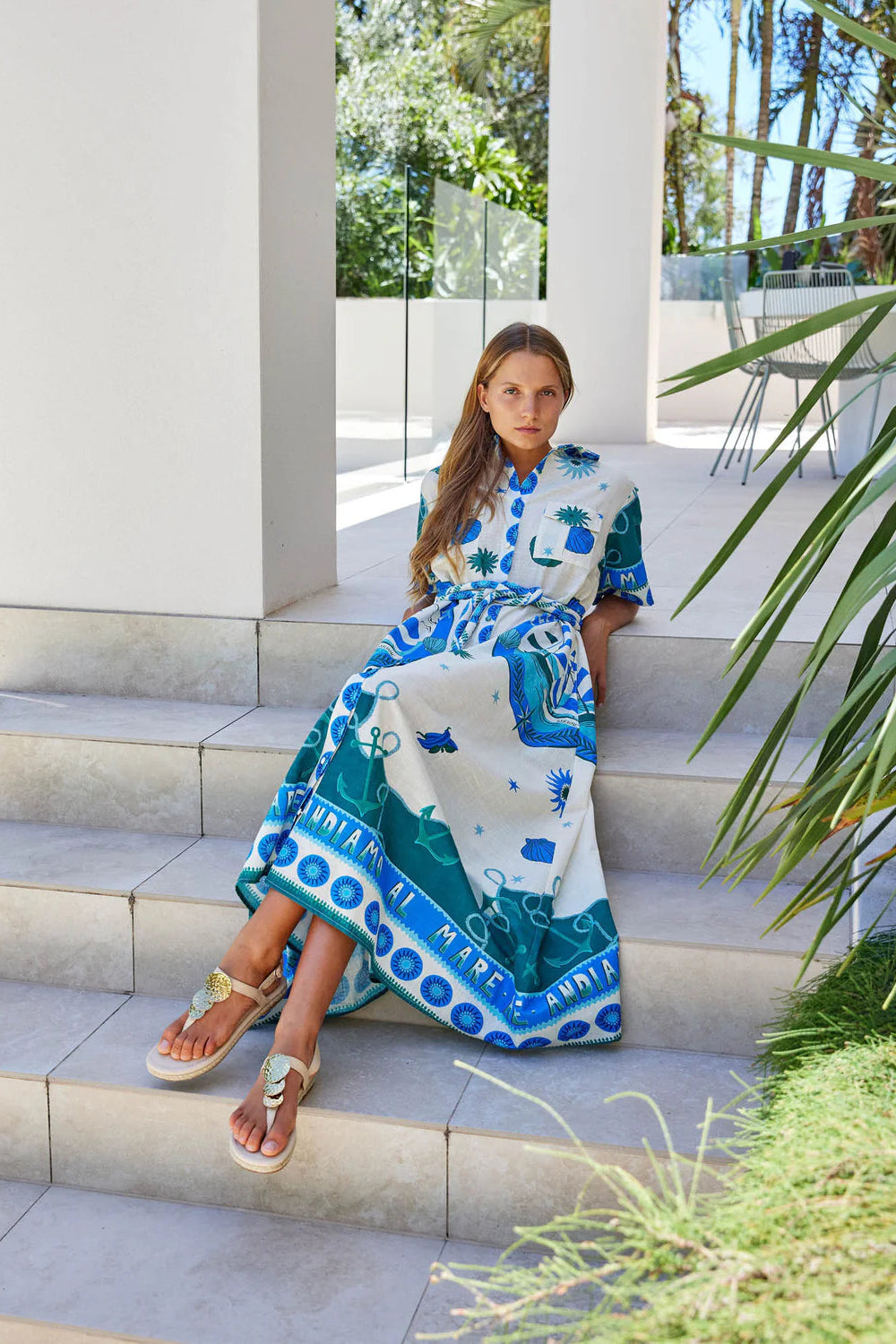 Woman in a patterned dress sitting on steps outdoors with greenery around.
