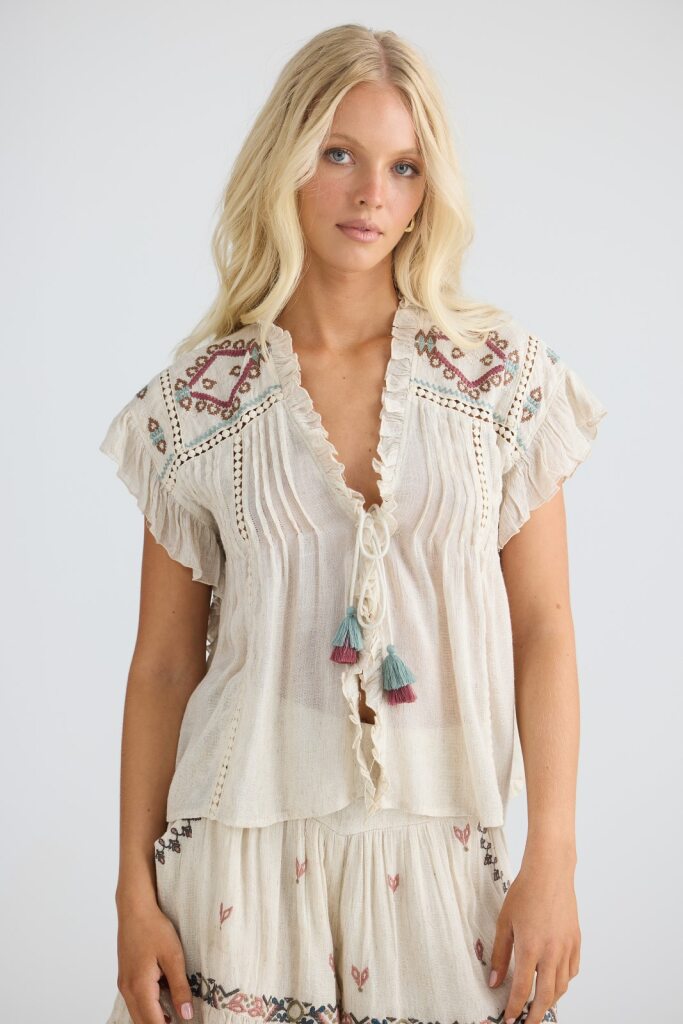 Woman wearing a white embroidered top with tassels on a plain background