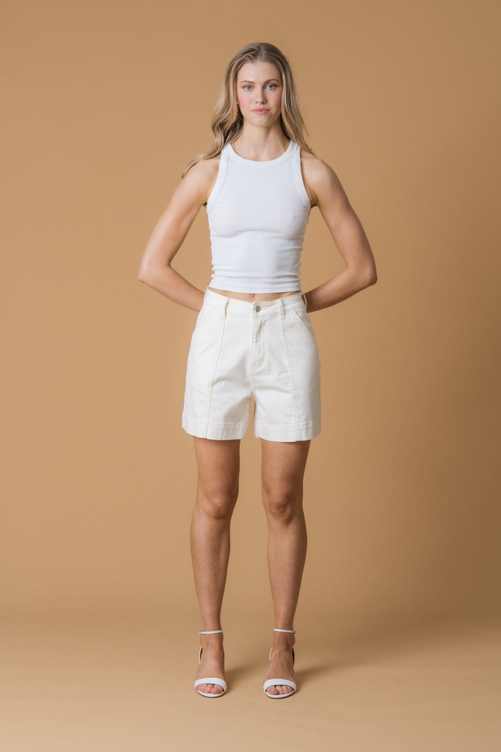 Woman wearing a white tank top and beige shorts against a brown background