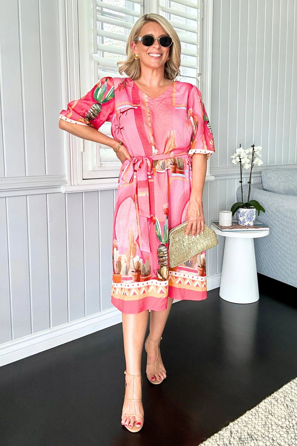 Woman in a pink dress with cactus print standing in a room with white walls and a gray sofa.