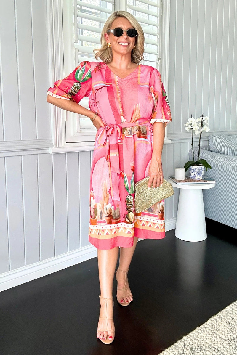 Woman in a pink dress with cactus print standing in a room with white walls and a gray sofa.