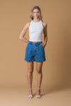 Woman wearing a white tank top and blue denim shorts against a brown background