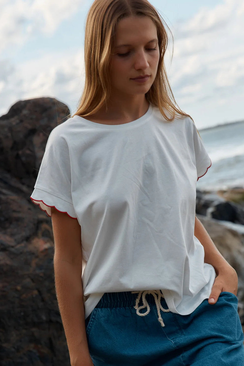 Woman wearing a white t-shirt with red trim and blue pants standing on a rocky beach.
