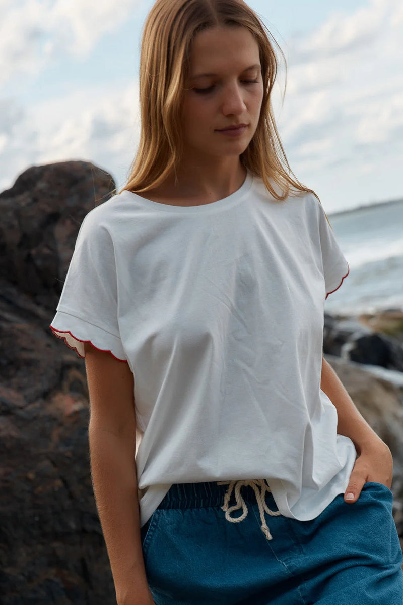 Woman wearing a white t-shirt with red trim and blue pants standing on a rocky beach.
