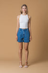 Woman wearing a white tank top and blue denim shorts against a beige background